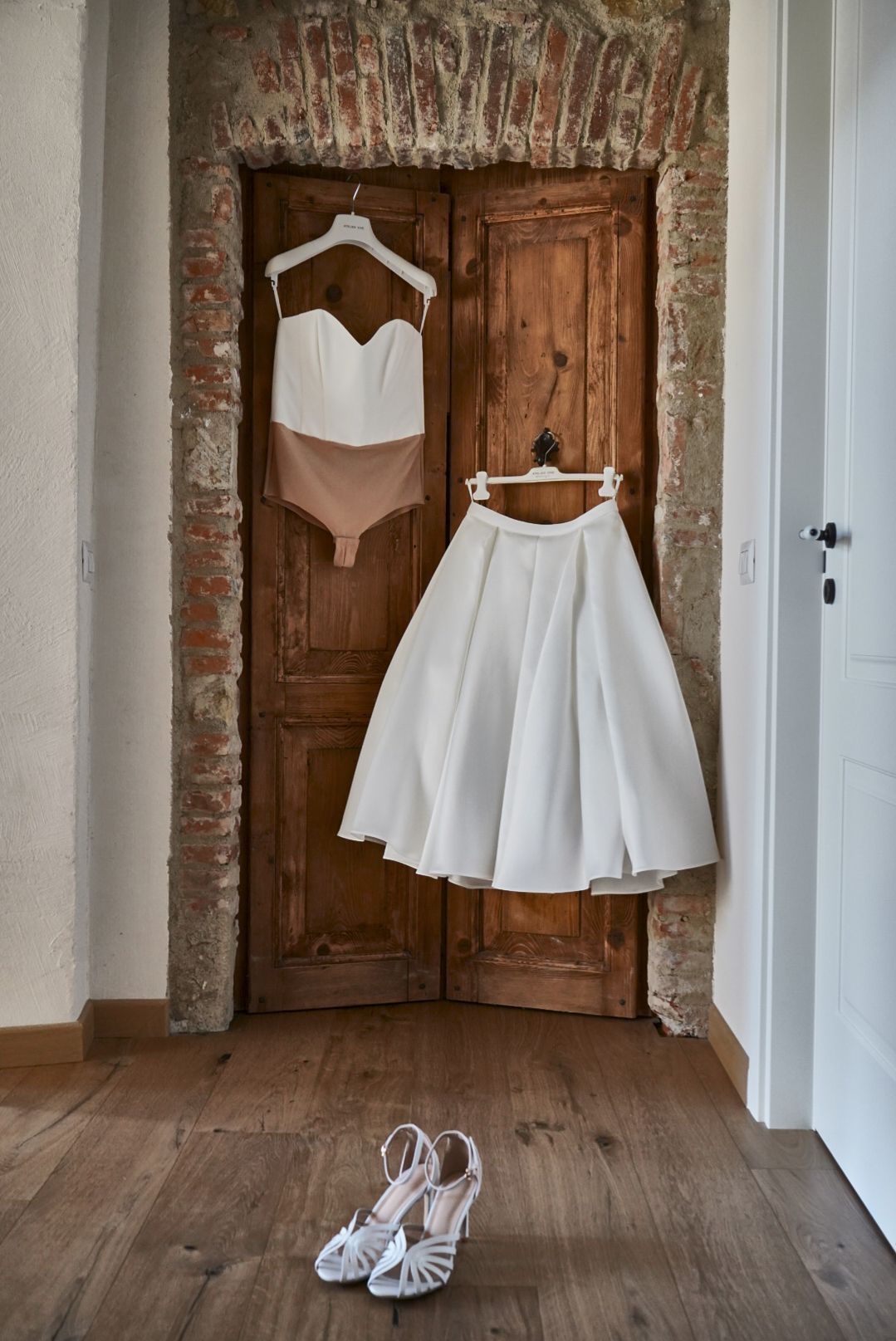 Abito da sposa appeso a una porta antica in legno, sandali bianchi sul parquet